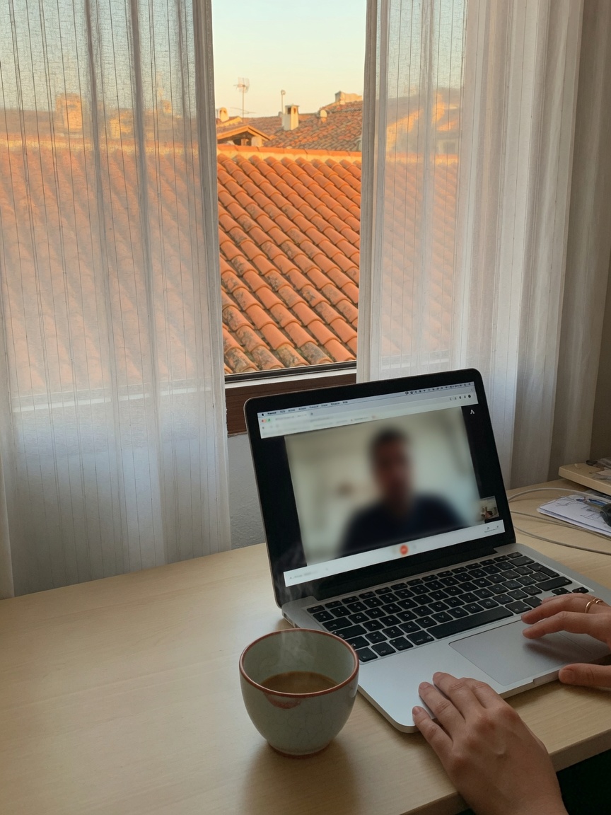 An online psychology session: a laptop by the window, a blurred figure on the screen, hands on the keyboard.